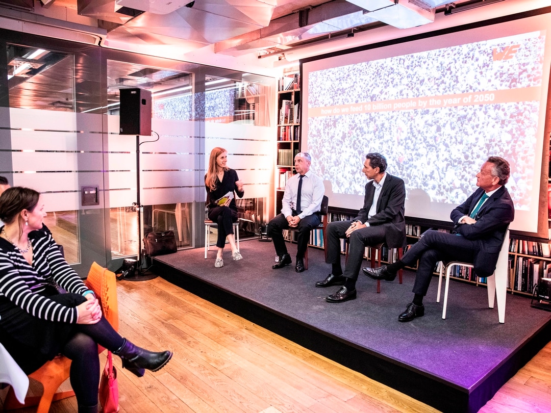 Four people sit on a podium and discuss. Two people from the audience are seen