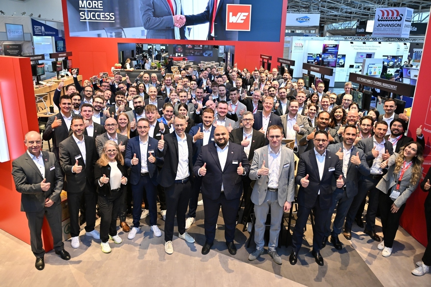 Team photo at the trade fair booth A large group of people poses at a trade fair booth and gives a thumbs-up to the camera