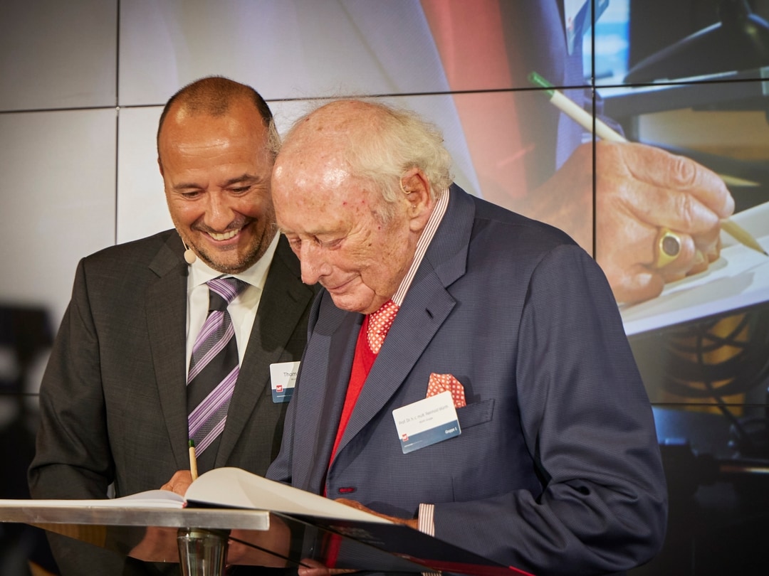 Auf dem Foto sind zwei Männer zu sehen. Links steht ein CEO und rechts ein bekannter Unternehmer. Beide schauen lächelnd auf das Pult. Der Unternehmer unterzeichnet ein Papier mit einem Bleistift.