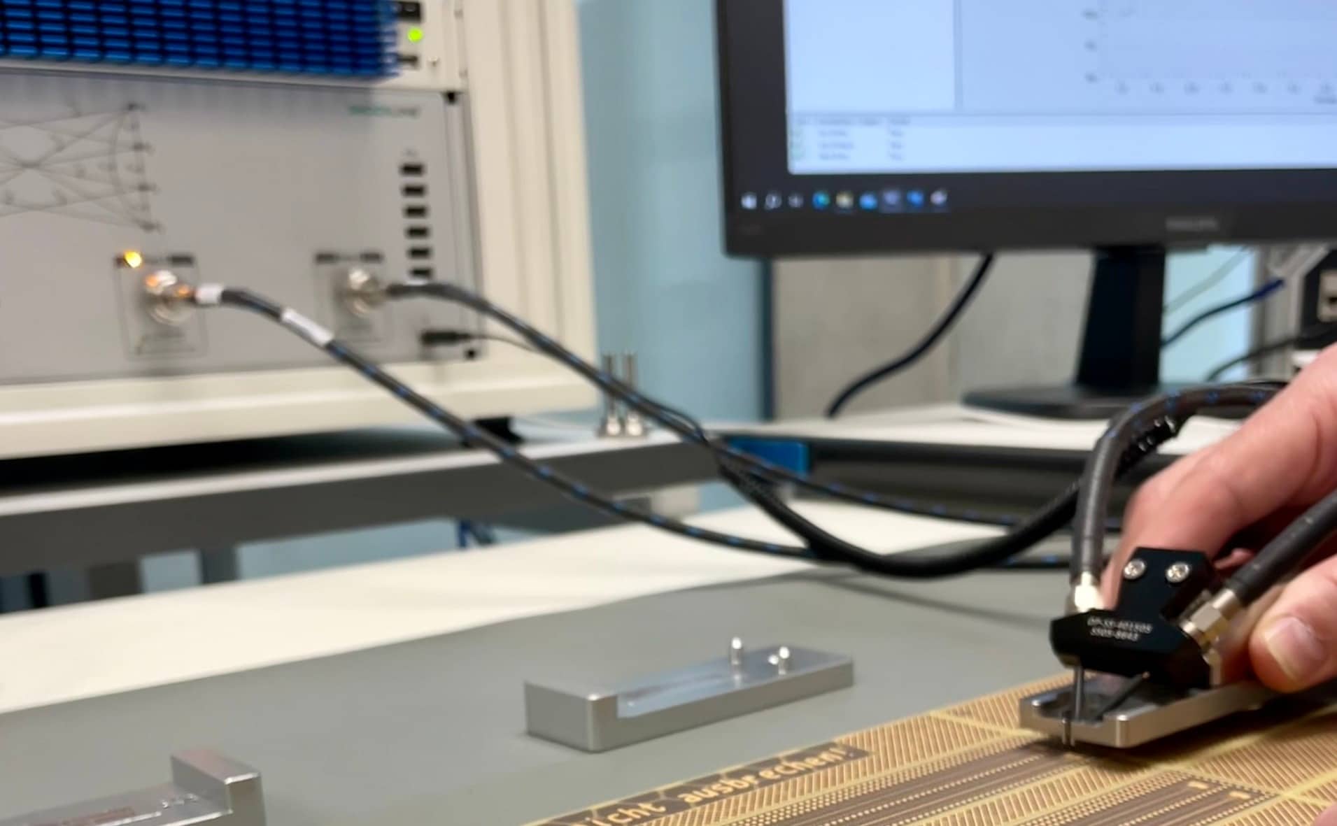 CBT WEdesign Polar Instrument EMV Test Eine Hand bedient ein Messgerät auf einer Leiterplatte während im Hintergrund das Testgerät und ein Bildschirm zu sehen sind