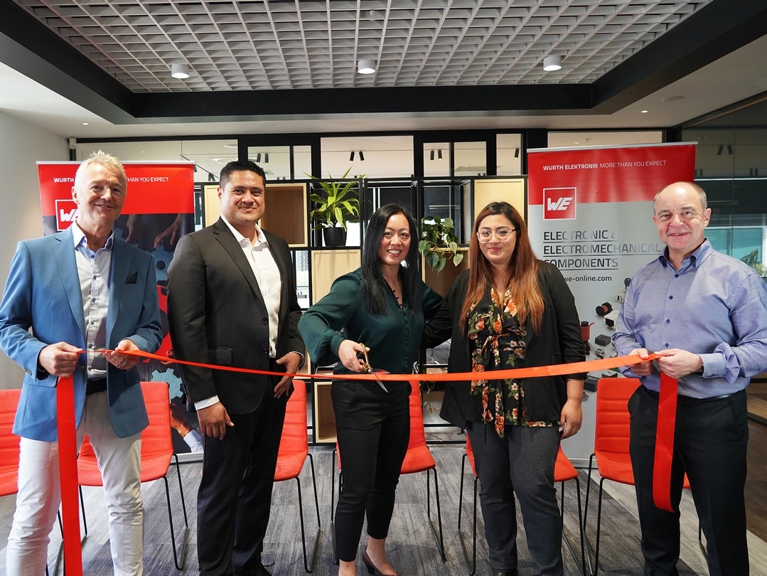 A group of people stand in front of a red ribbon in an office. The female person in the middle cuts a red ribbon. Everyone is smiling.