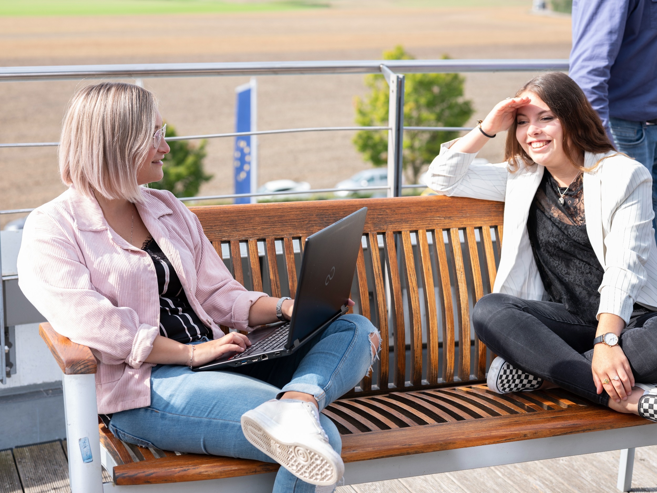 bachelor program Two women are sitting on a bench chatting, one of them has a laptop on her lap. The two of them are outside on a terrace