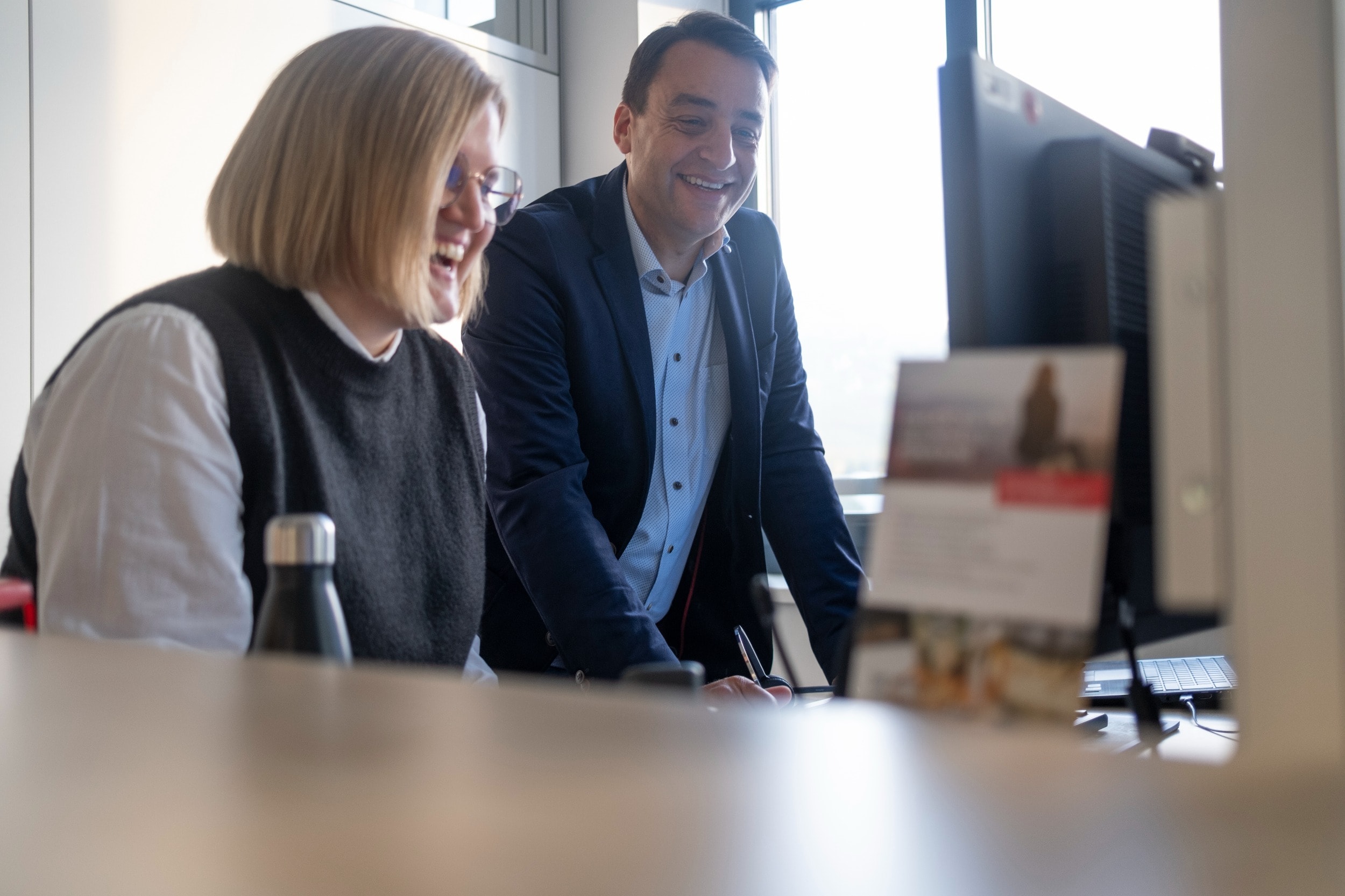 FAQs Bewerbungsprozess Mann und Frau stehen nebeneinander lachend vor einem Bildschirm in einem Büro