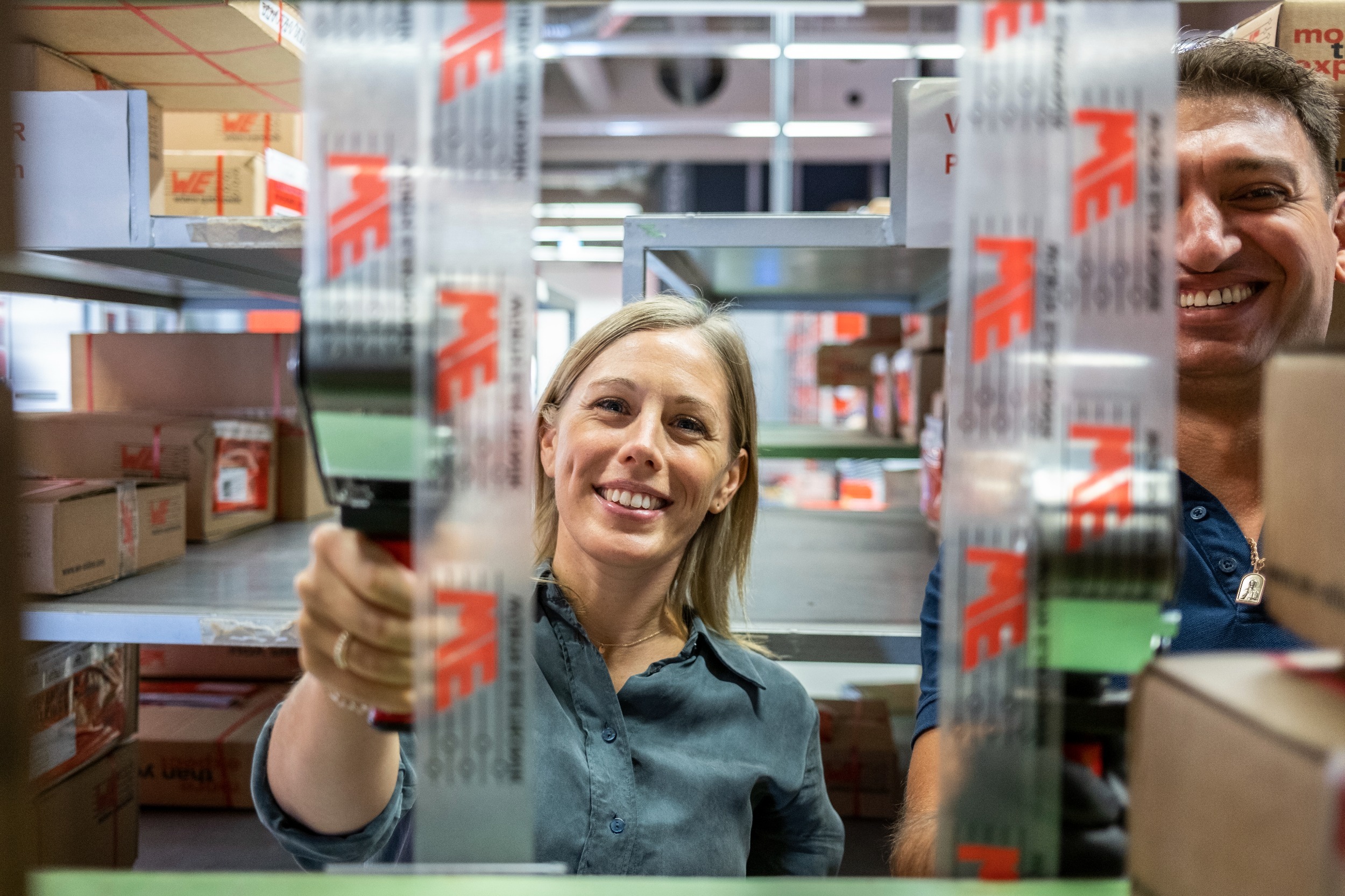 Man and woman in warehouse with label device A woman and a man are smiling in front of a shelf with a labelling device, adhesive tape comes out of the labelling device which is transparent and has the Würth Elektronik logo on it.