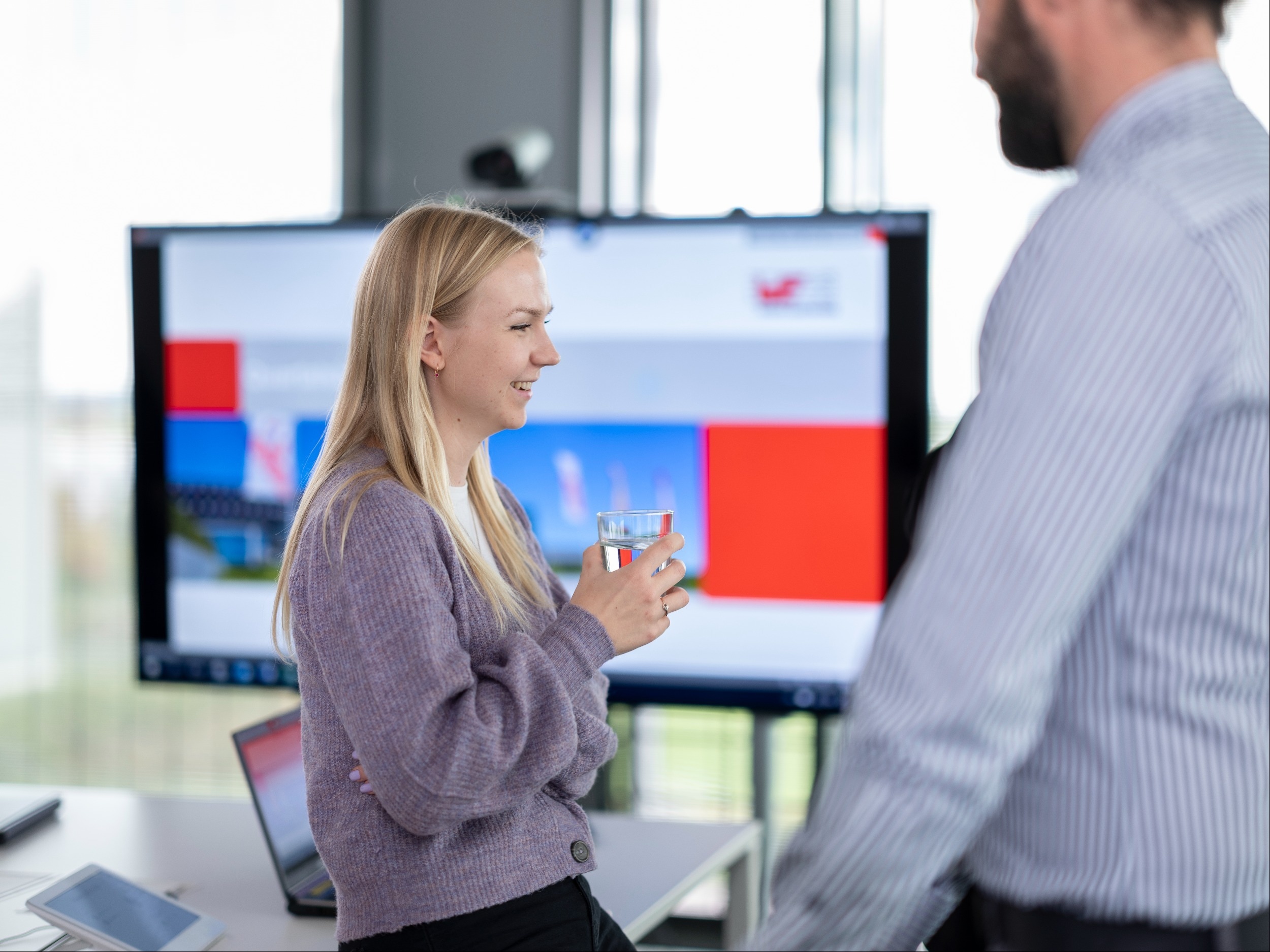 Woman in meeting room A woman with a glass of water in her hand leans against a table, in front of her stands a man. In the background is a monitor