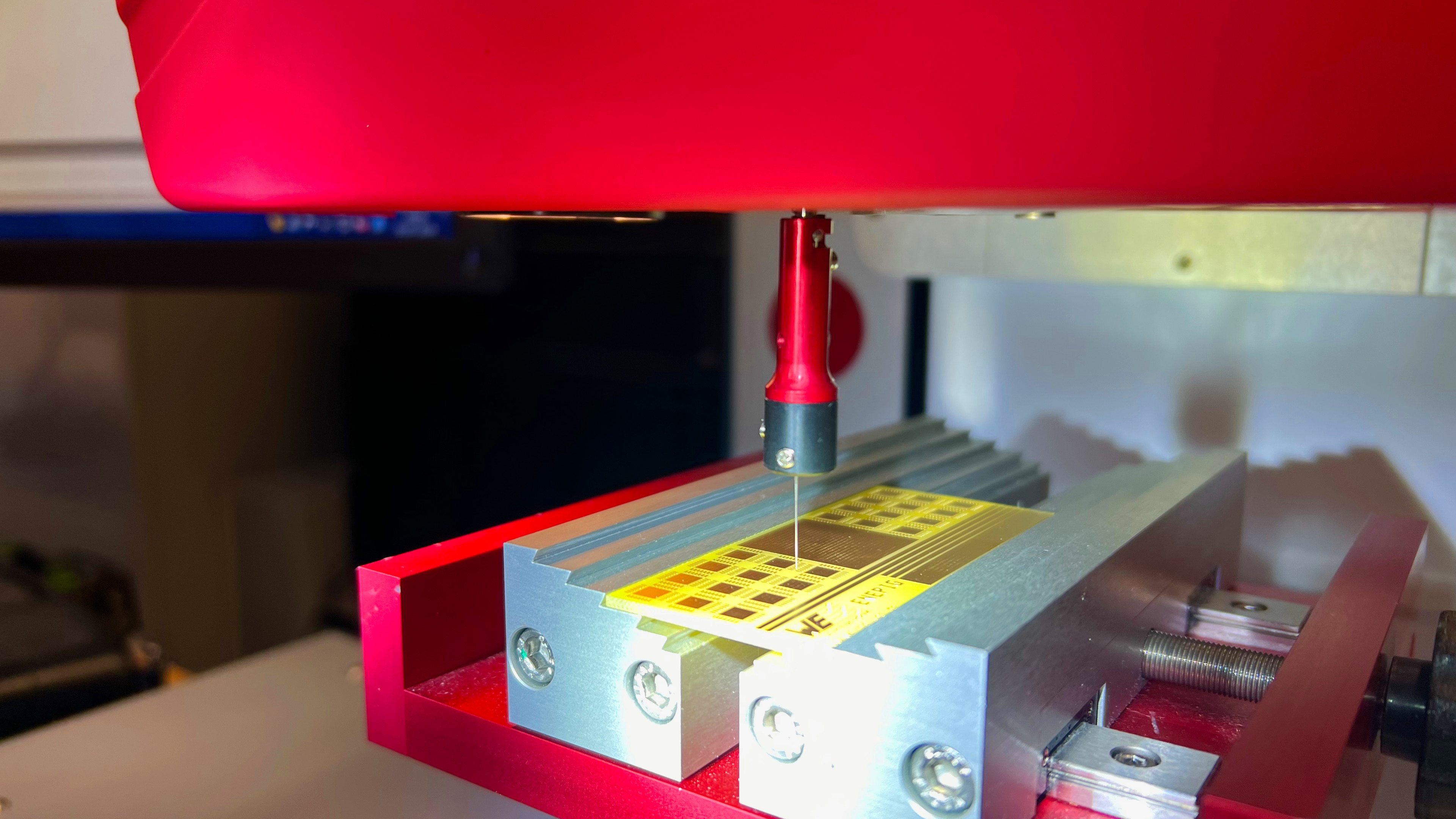 Eine Nahaufnahme einer Metallprüfmaschine in einem Labor mit einer Metallprobe, die von einer roten Klemme gehalten wird. Die Einrichtung scheint für Abzieh- oder Schertests zu dienen.